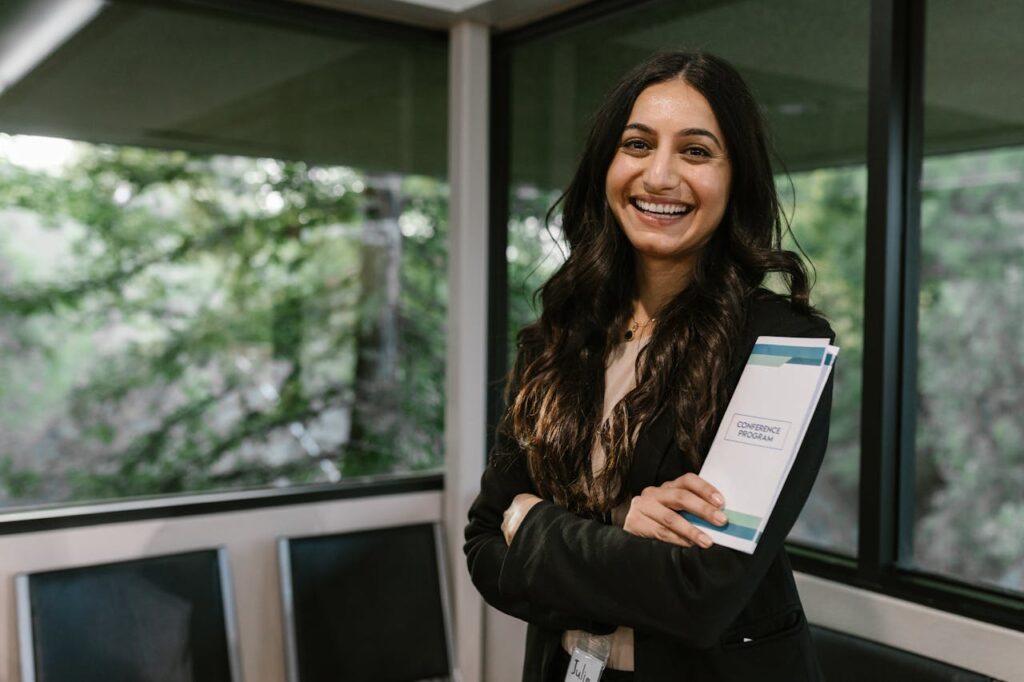 pexels-photo-7648319 Confident woman holding conference program, smiling in modern indoor setting.