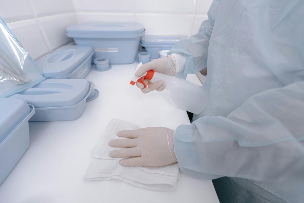 A healthcare worker wearing gloves sanitizes a surface with a spray bottle in a medical setting.
